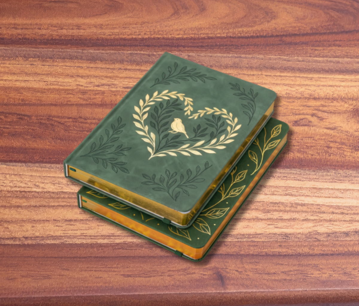 A stack of two decorative green hardcover books with gold leaf detailing, including a heart-shaped wreath design on the top book, displayed on a wooden tabletop against a pink background.