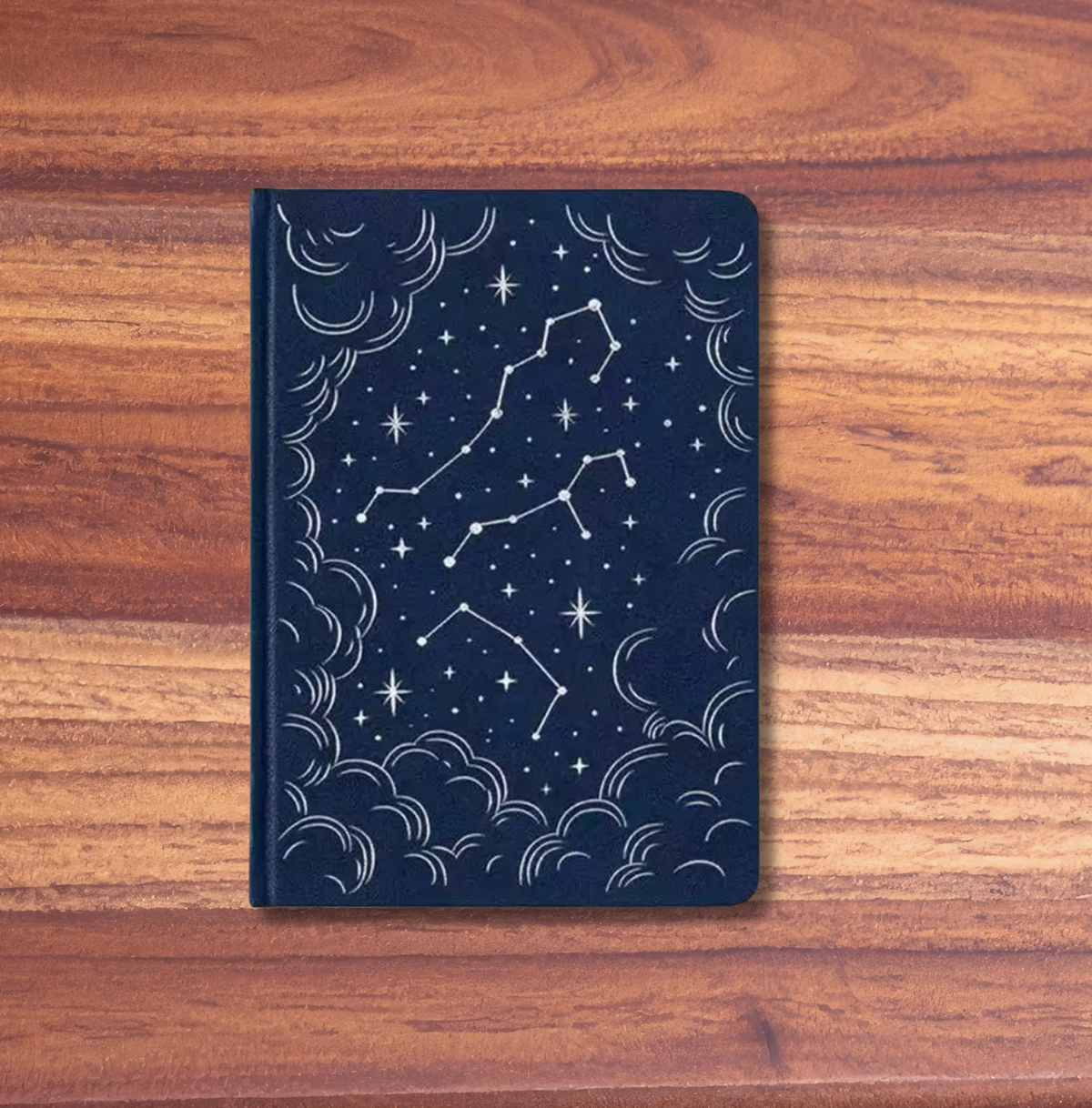 A dark navy blue notebook with a white celestial pattern of stars and constellations, resting on a wooden surface with a pink background.