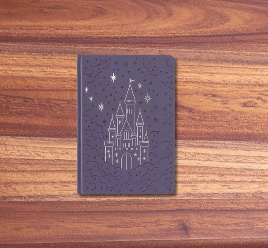 A dark purple notebook with a white line-art illustration of a castle under stars, displayed on a wooden surface with a pink background.