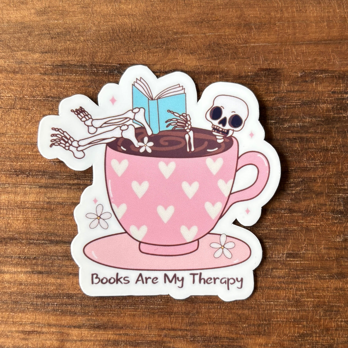 Sticker of a pink teacup with skeletons and a book, on a wooden surface.