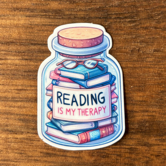 Sticker of a jar with books and glasses, featuring 'Reading is my Therapy' text on a wooden surface.