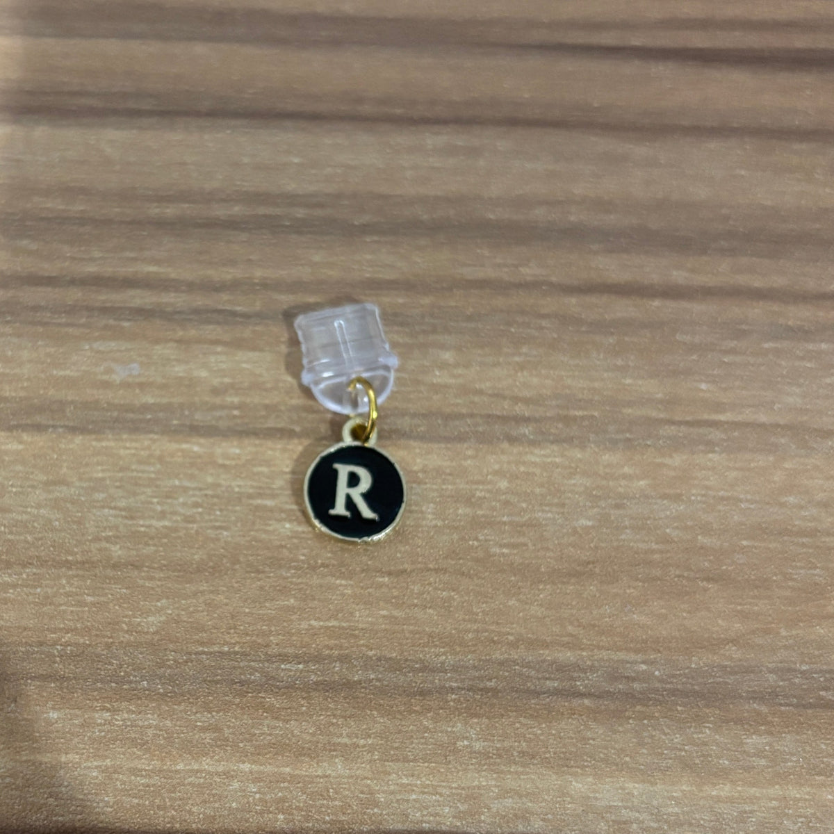 A small circular black charm pendant with a gold border and the letter 'R' in white, hanging from a clear plastic display stand on a wooden surface.