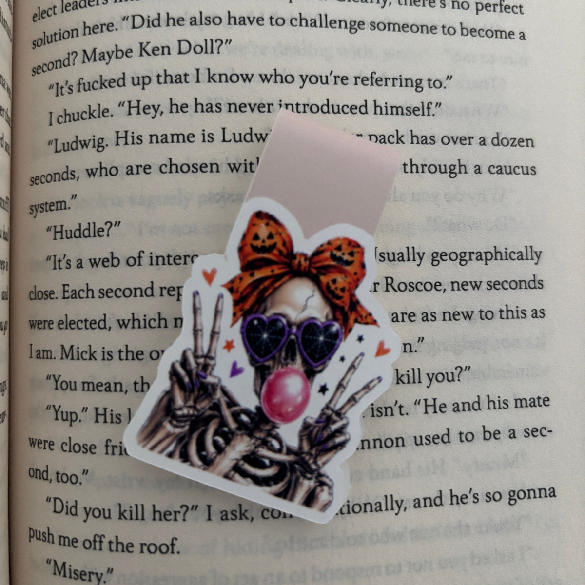 Skeleton Magnetic Bookmark, skeleton has pumpkin hairbow, blowing a bubble gum bubble and this giving peace signs on a book