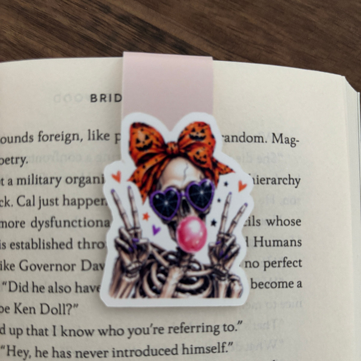 A magnetic bookmark featuring a cartoon character skeleton with a lpumpkin-print bow, heart-shaped sunglasses, and a pink bubblegum bubble, shown clipped over an open book page.