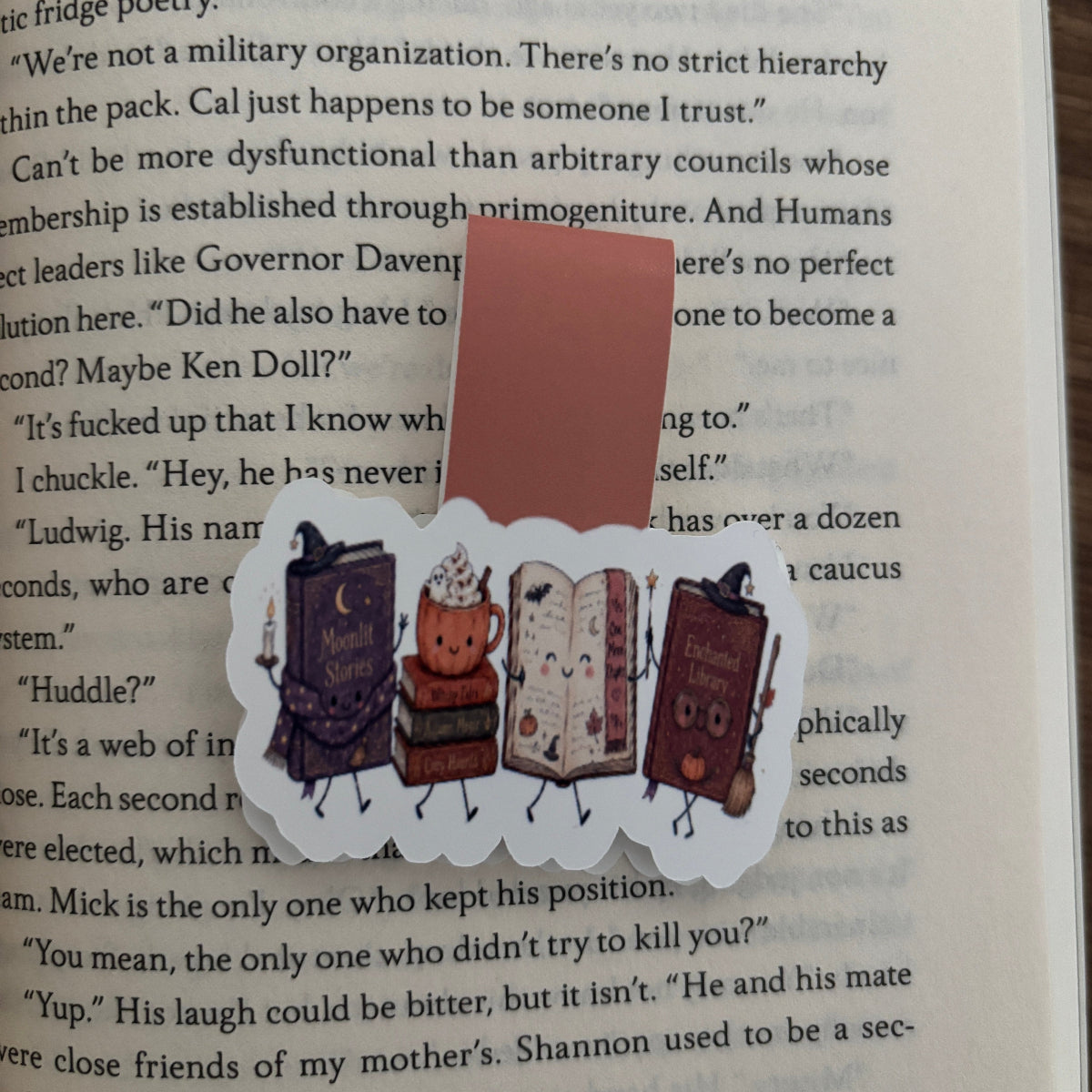 A die-cut magnetic bookmarkvfeaturing four cartoon books with faces and legs in the theme of halloween, placed on top of an open book page.
