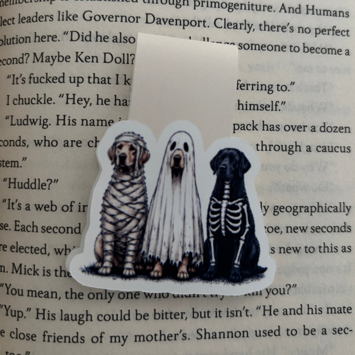 A die-cut magnetic bookmark featuring three dogs dressed in Halloween costumes, placed on top of an open book page. The left dog is wrapped in bandages, the middle dog is in a white ghost costume, and the right dog has a black skeleton outfit.