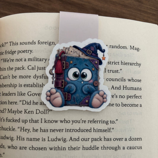 A die-cut magnetic bookmark of a cute blue monster sitting on an open book, photographed on a wooden surface with book pages visible in the background.