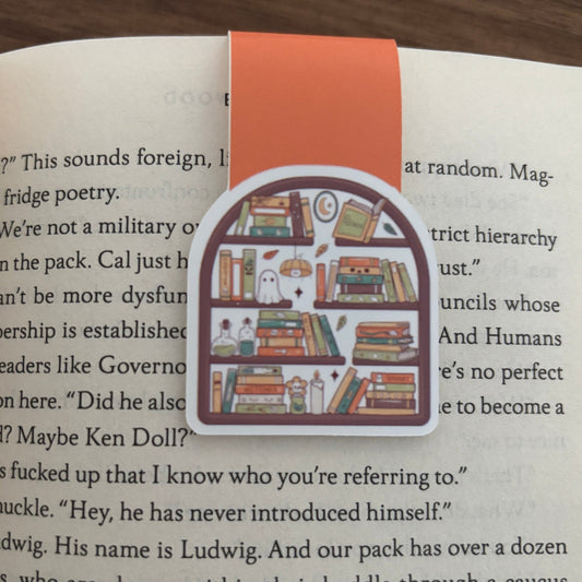 A die-cut magnetic bookmark shaped like a bookshelf with books, a owl, and small illustrations, attached to an orange bookmark clip on an open book.
