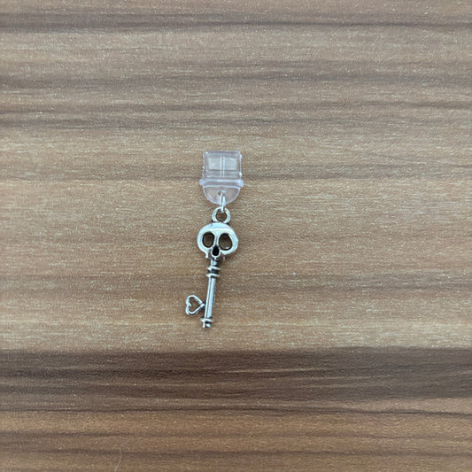 A silver-toned key-shaped charm with a skull detail at the top, attached to a clear plastic hanger, placed on a brown wood-grain surface.