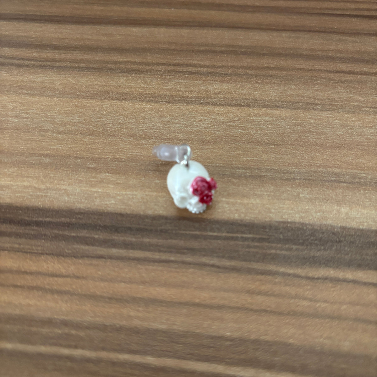 A small skull-shaped pendant or charm with a red flower accent and a clear bead at the top, resting on a brown wood-grain surface.