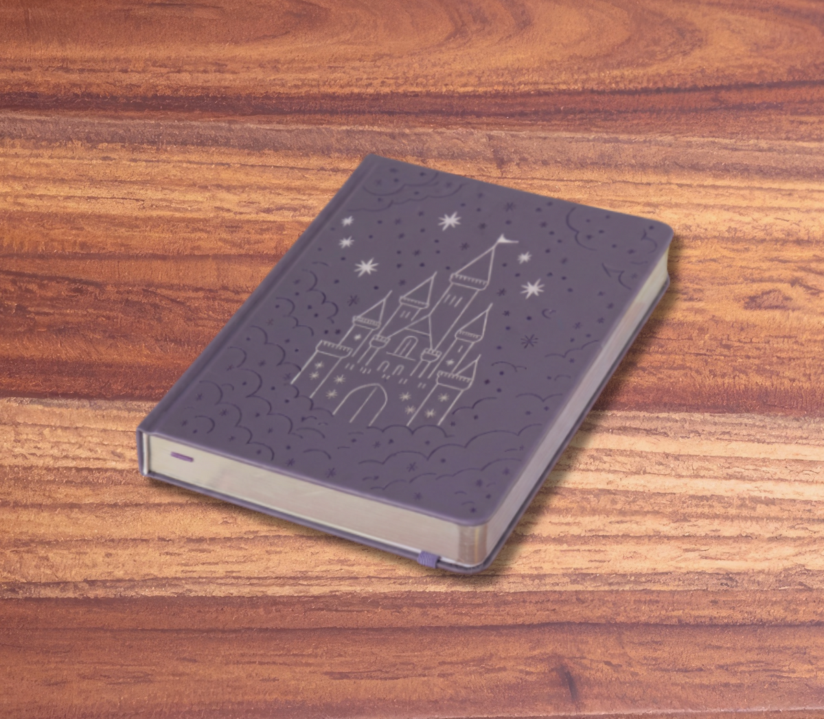 A dark purple hardcover notebook with an embossed castle and stars design, resting on a wooden tabletop against a solid pink background.