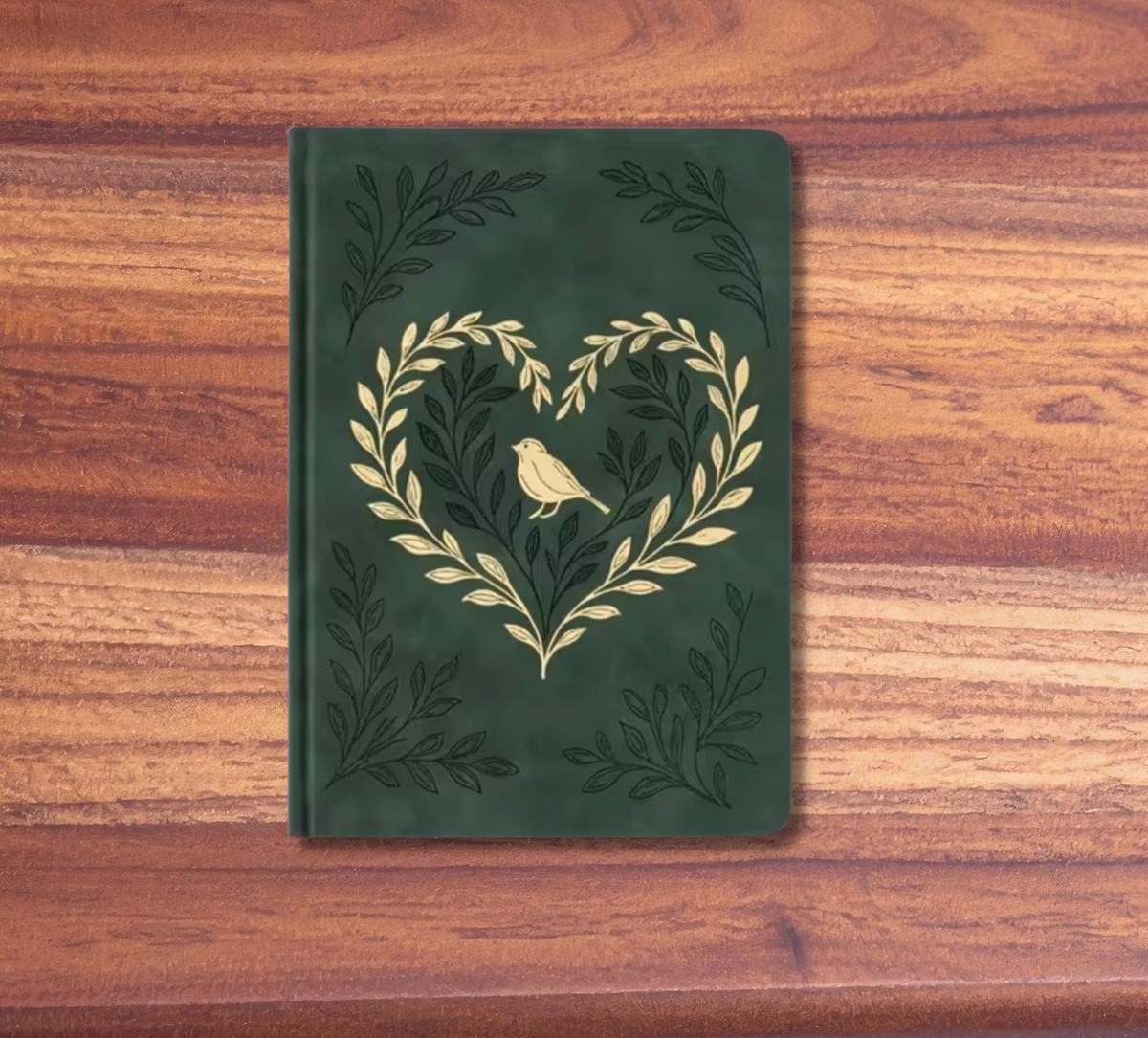A dark green hardcover notebook with an embossed gold laurel wreath and bird design, displayed on a wooden surface with a pink background.
