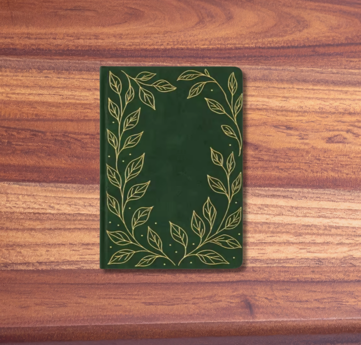 A dark green notebook with a gold leaf border design, placed on a wooden surface with pink background borders.