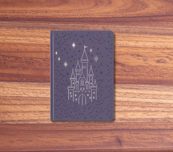 A dark purple hardcover notebook featuring a white line-art illustration of a castle under stars, displayed on a wooden surface with a pink background.