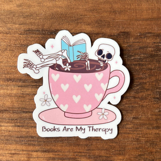 Sticker of a pink teacup with skeletons and a book, on a wooden surface.