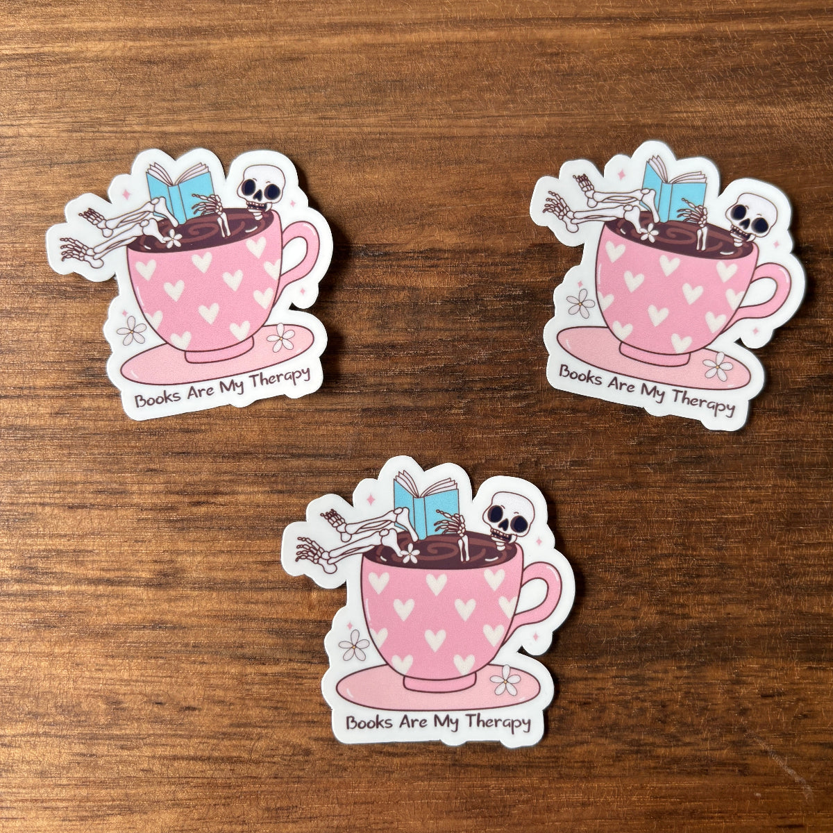 Three stickers of a pink teacup with books and skeletons on a wooden surface.