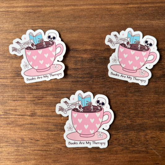 Three stickers of a pink teacup with books and skeletons on a wooden surface.
