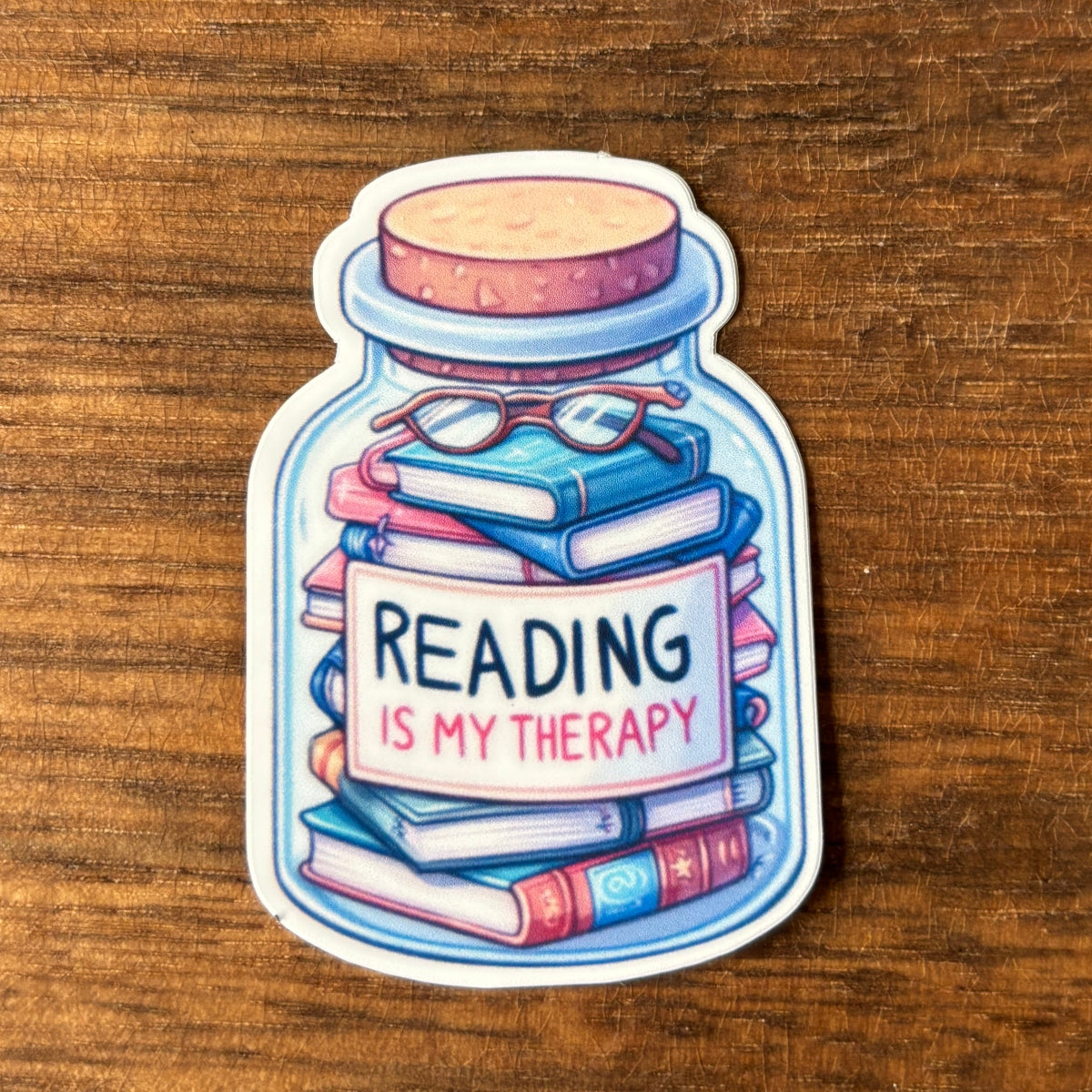 Sticker of a jar with books and glasses, featuring 'Reading is my Therapy' text on a wooden surface.