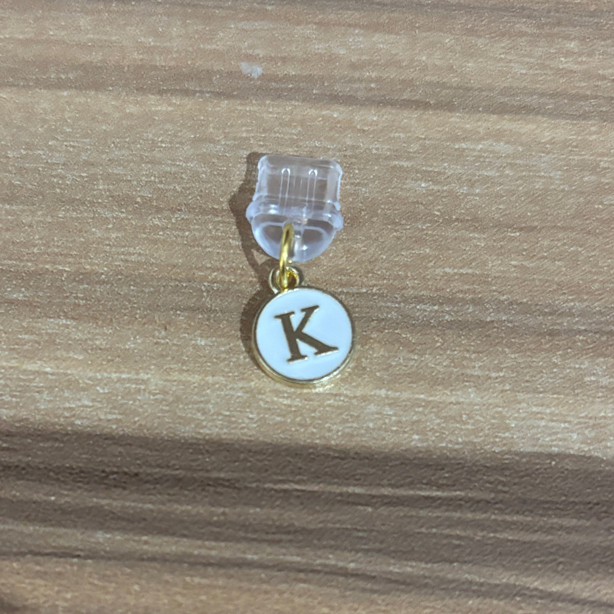 A small initial charm featuring a gold-tone circular pendant with a white enamel background and a dark brown letter 'K', suspended from a clear plastic clip on a wooden surface.