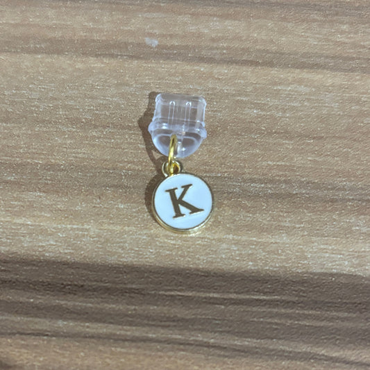A small initial charm featuring a gold-tone circular pendant with a white enamel background and a dark brown letter 'K', suspended from a clear plastic clip on a wooden surface.