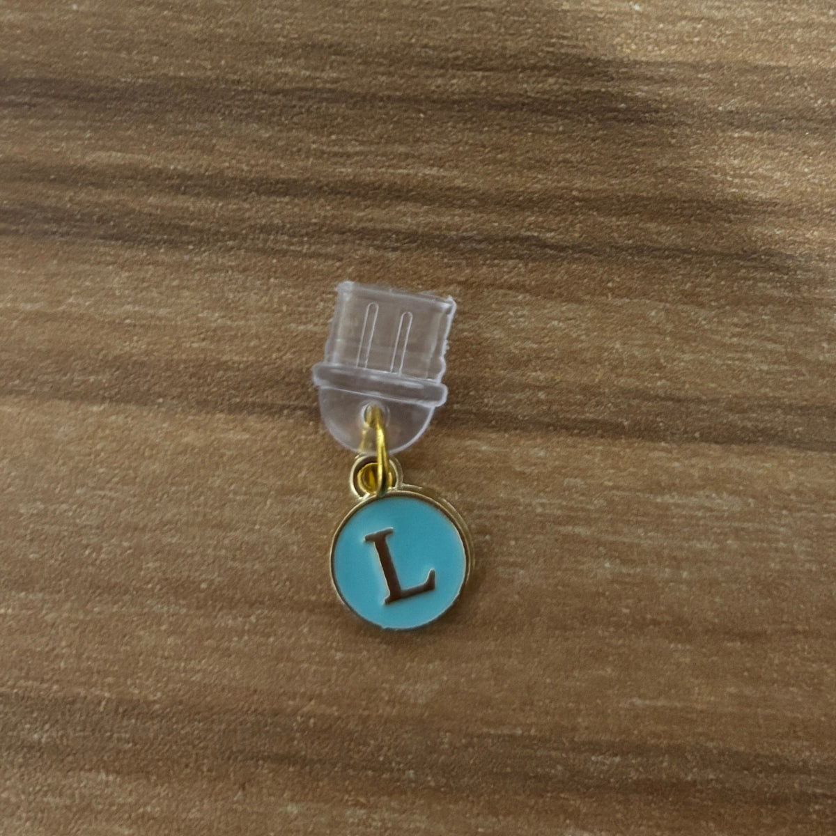 A small round initial charm with a turquoise blue enamel face and a gold-tone letter 'L', attached to a clear plastic display stand on a brown wood-grain surface.