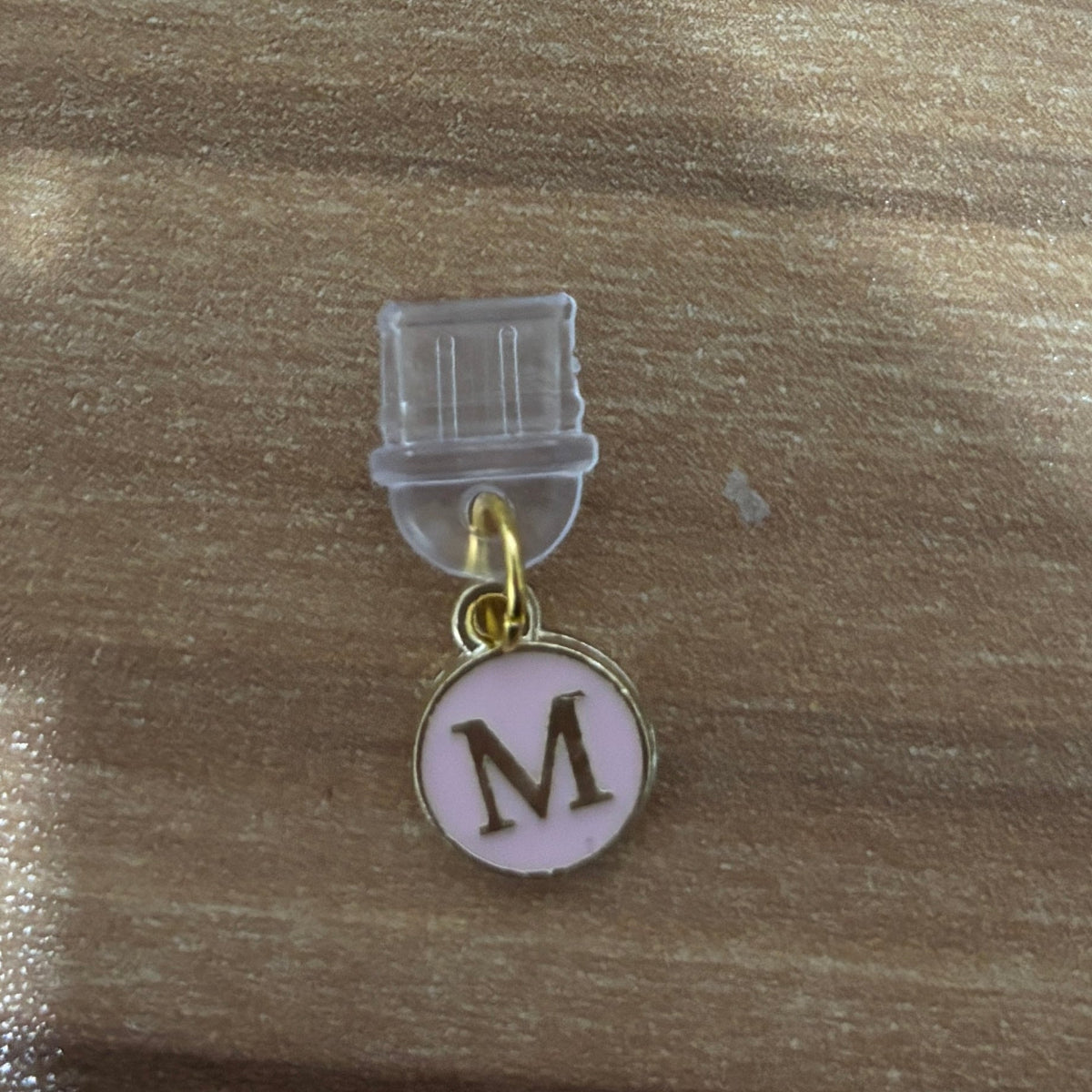 A small personalized charm with a circular light pink enamel face featuring a gold metal border and a dark brown letter 'M'. The charm is suspended from a gold ring and attached to a clear plastic clip, shown on a wooden surface.