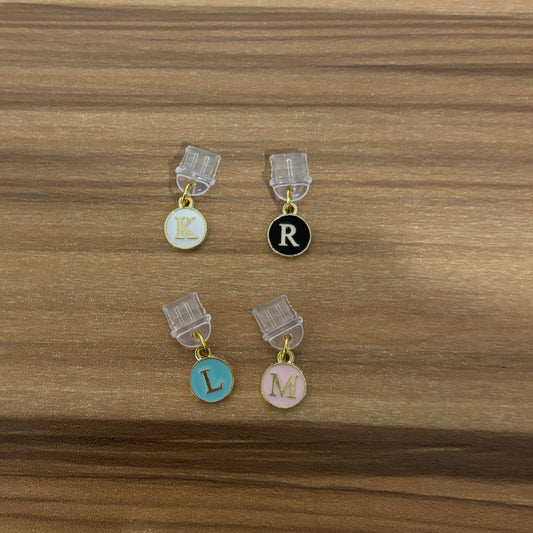 Four initial letter charms with clear plastic clips, displayed on a wooden surface. The letters are K, R, L, and M in gold-tone font on white, black, light blue, and light pink circular charms.