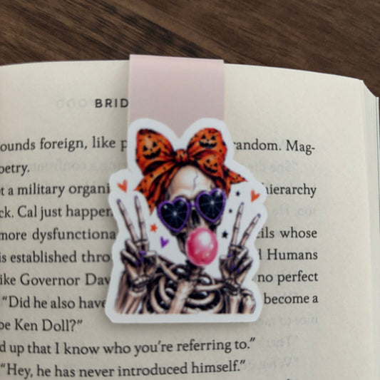 A magnetic bookmark featuring a cartoon character skeleton with a lpumpkin-print bow, heart-shaped sunglasses, and a pink bubblegum bubble, shown clipped over an open book page.