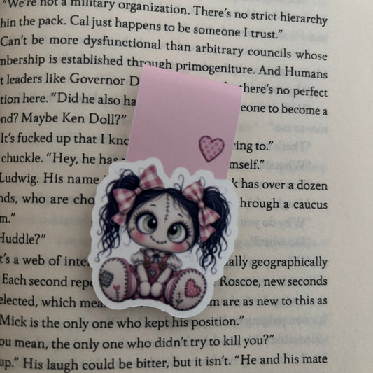 A die-cut magnetic bookmark featuring a cartoon girl with dark hair, a pink bow, and a pink heart, placed on top of an open book page with printed text.