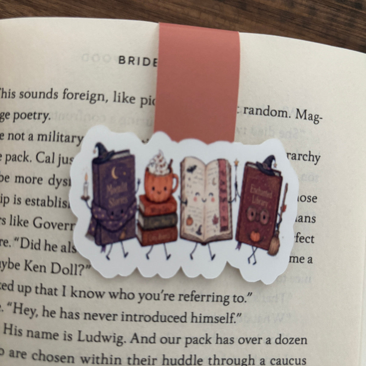 A cute die-cut mangetic bookmark featuring anthropomorphic books with faces and limbs, themed for halloween resting on an open book page.