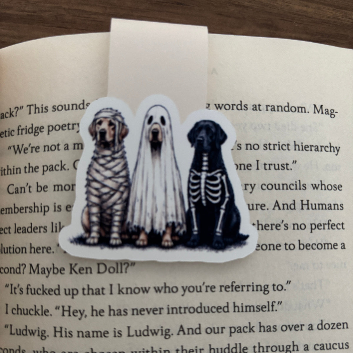 A die-cut magnetic bookmark featuring three dogs dressed as mummy, bride, and skeleton, placed on an open book page.