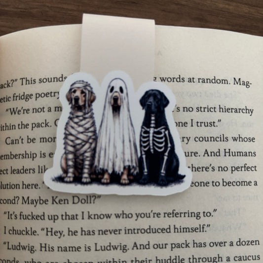 A die-cut magnetic bookmark featuring three dogs dressed as mummy, bride, and skeleton, placed on an open book page.