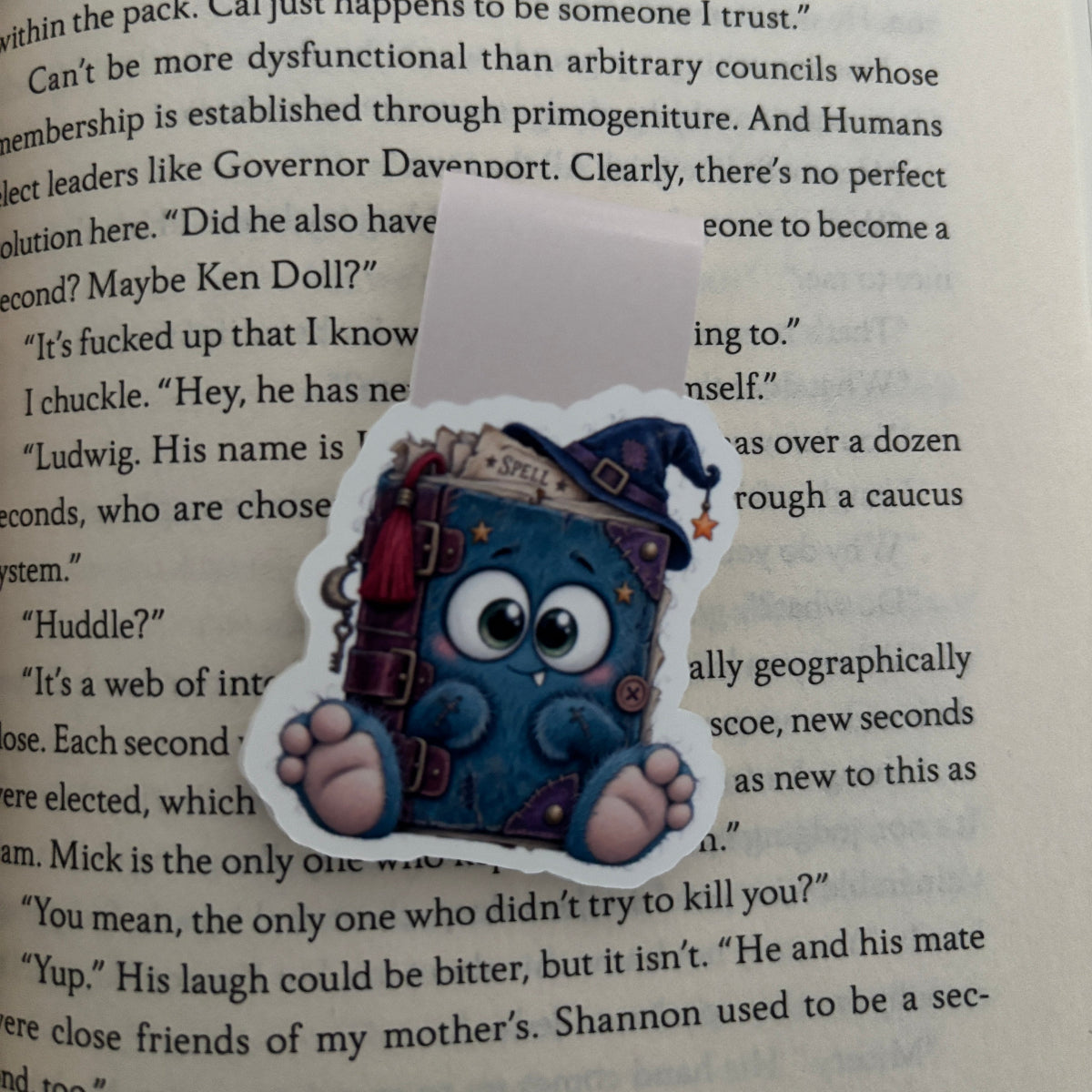 A die-cut magnetic bookmark featuring a cute blue cartoon character holding a stack of books, placed on top of a printed book page.