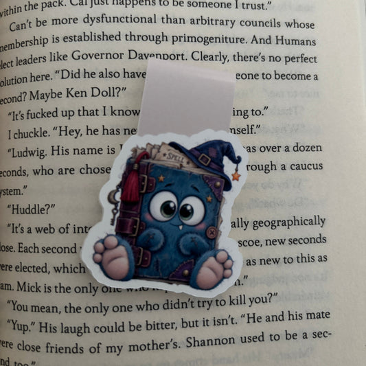 A die-cut magnetic bookmark featuring a cute blue cartoon character holding a stack of books, placed on top of a printed book page.