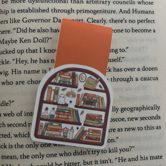 A die-cut magnetic bookmark featuring a whimsical bookshelf illustration with a ghost and small characters, placed on top of an open book page.