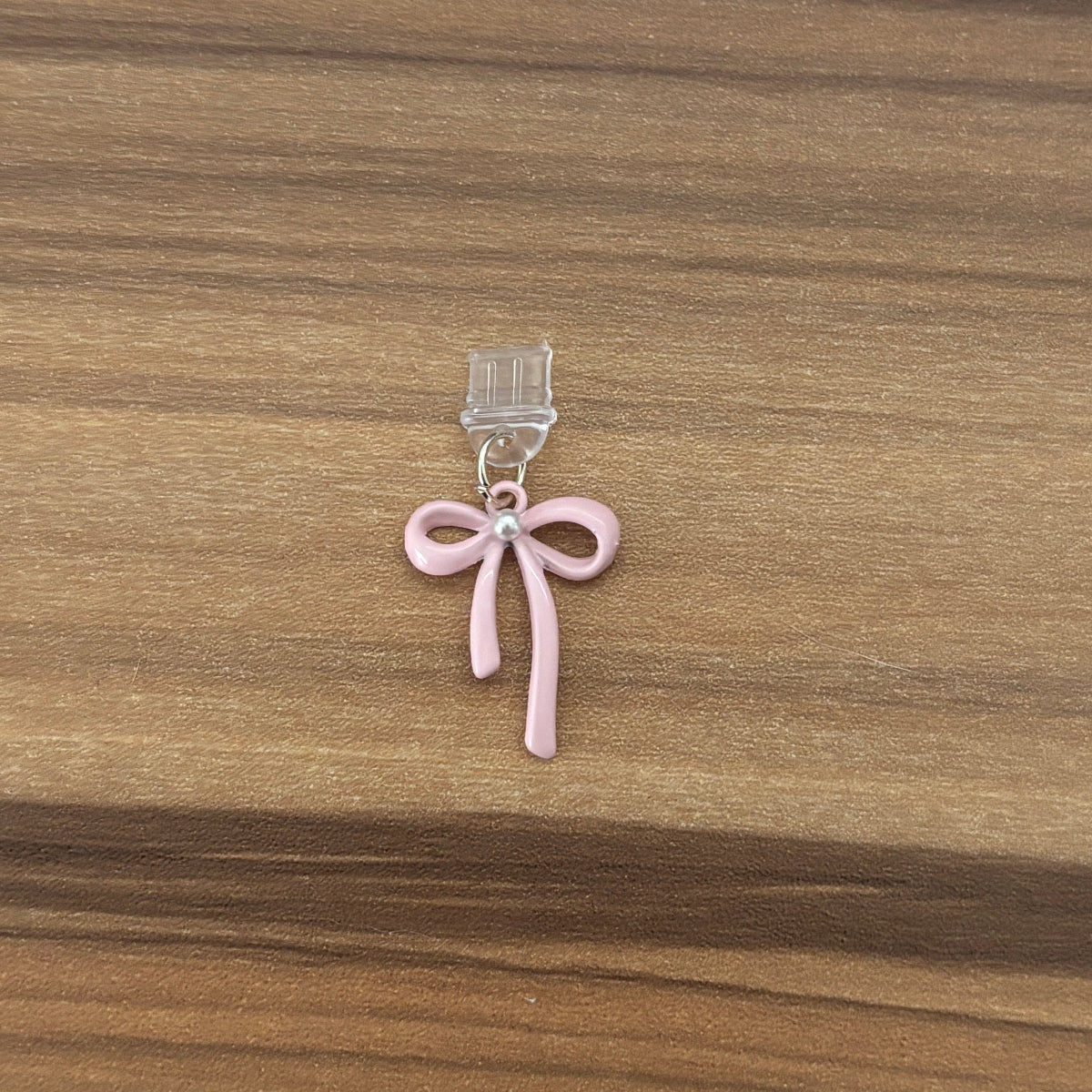 A small pink bow charm with a clear plastic clip at the top, resting on a brown wooden surface.