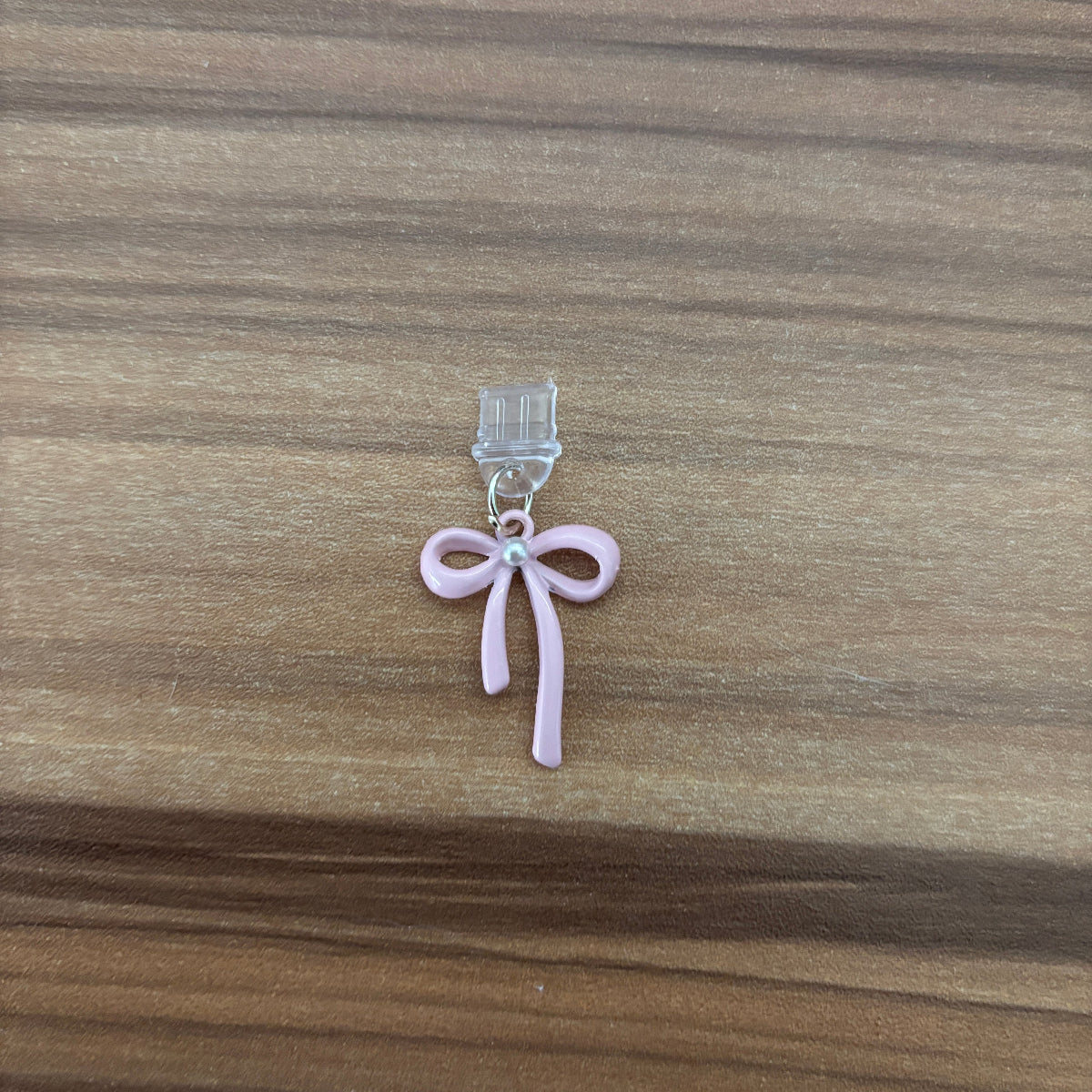 A small pink bow-shaped charm with a clear plastic clip, placed on a brown wooden surface.