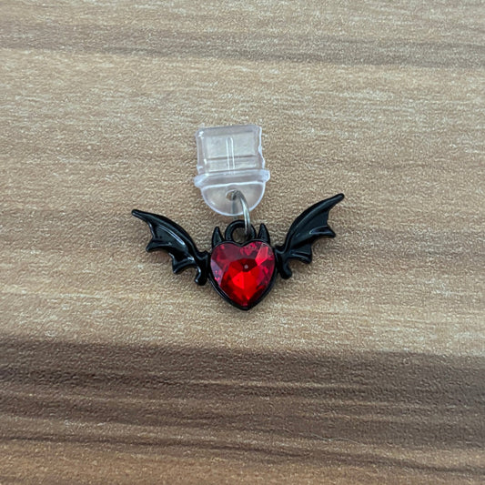 A small gothic-style accessory featuring a red faceted heart gem with black bat wings, attached to a clear plastic hook, displayed on a brown wood-grain surface.