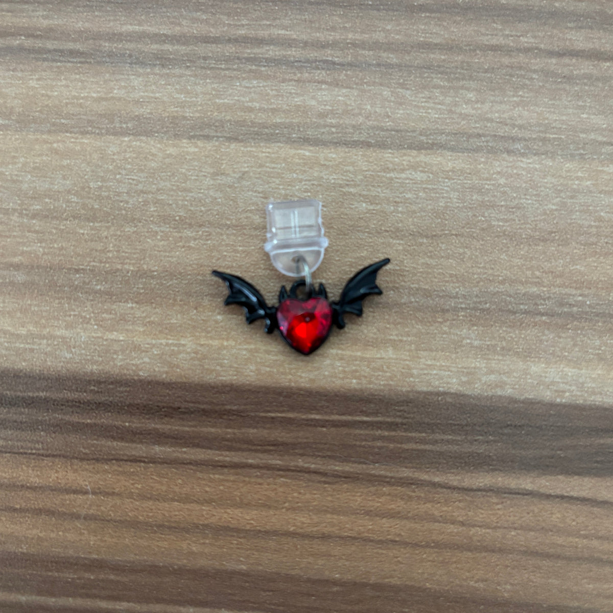 A small gothic-style charm featuring a red heart with black bat wings, attached to a clear plastic loop, shown on a wooden surface.