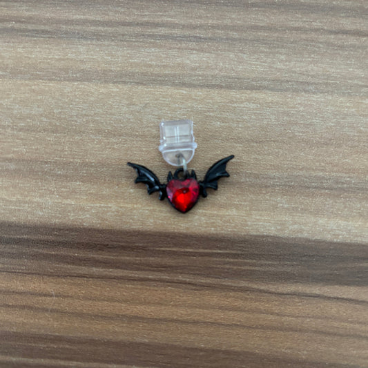 A small gothic-style charm featuring a red heart with black bat wings, attached to a clear plastic loop, shown on a wooden surface.