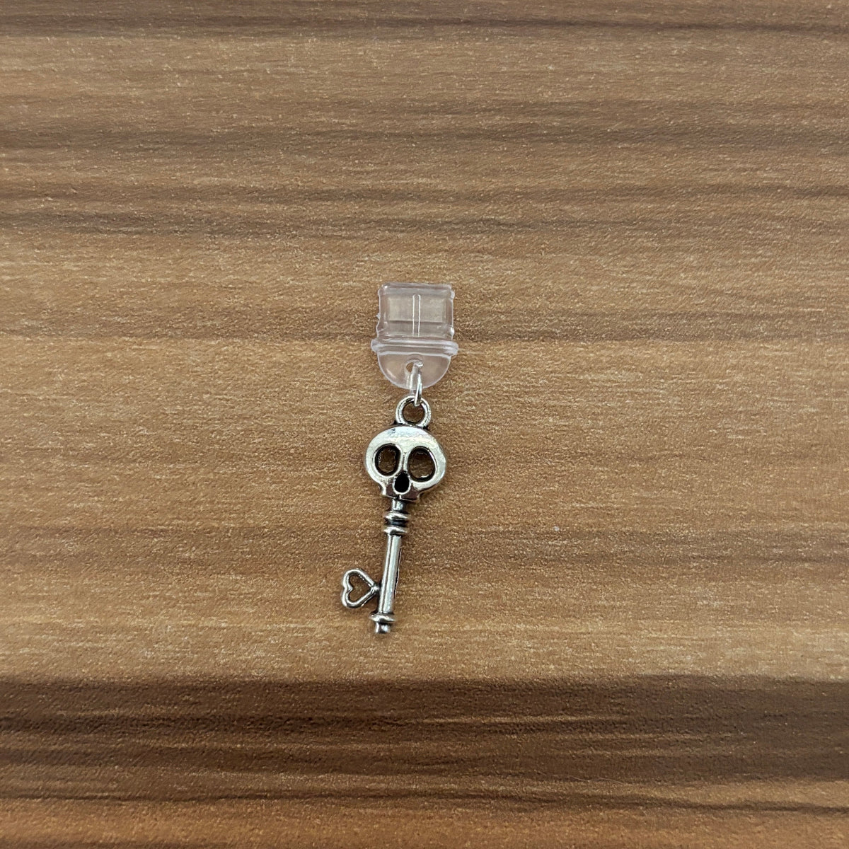 A silver-toned skull and key charm with a clear plastic hanger, displayed on a brown wooden surface.