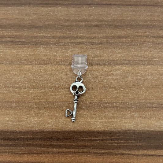 A silver-toned skull and key charm with a clear plastic hanger, displayed on a brown wooden surface.