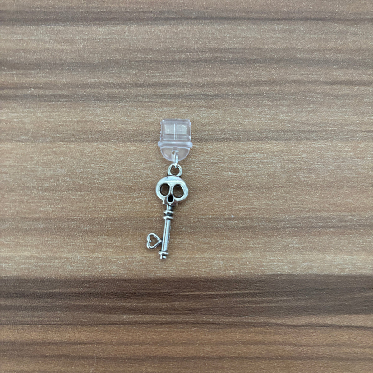A silver-toned key-shaped charm with a skull detail at the top, attached to a clear plastic hanger, placed on a brown wood-grain surface.
