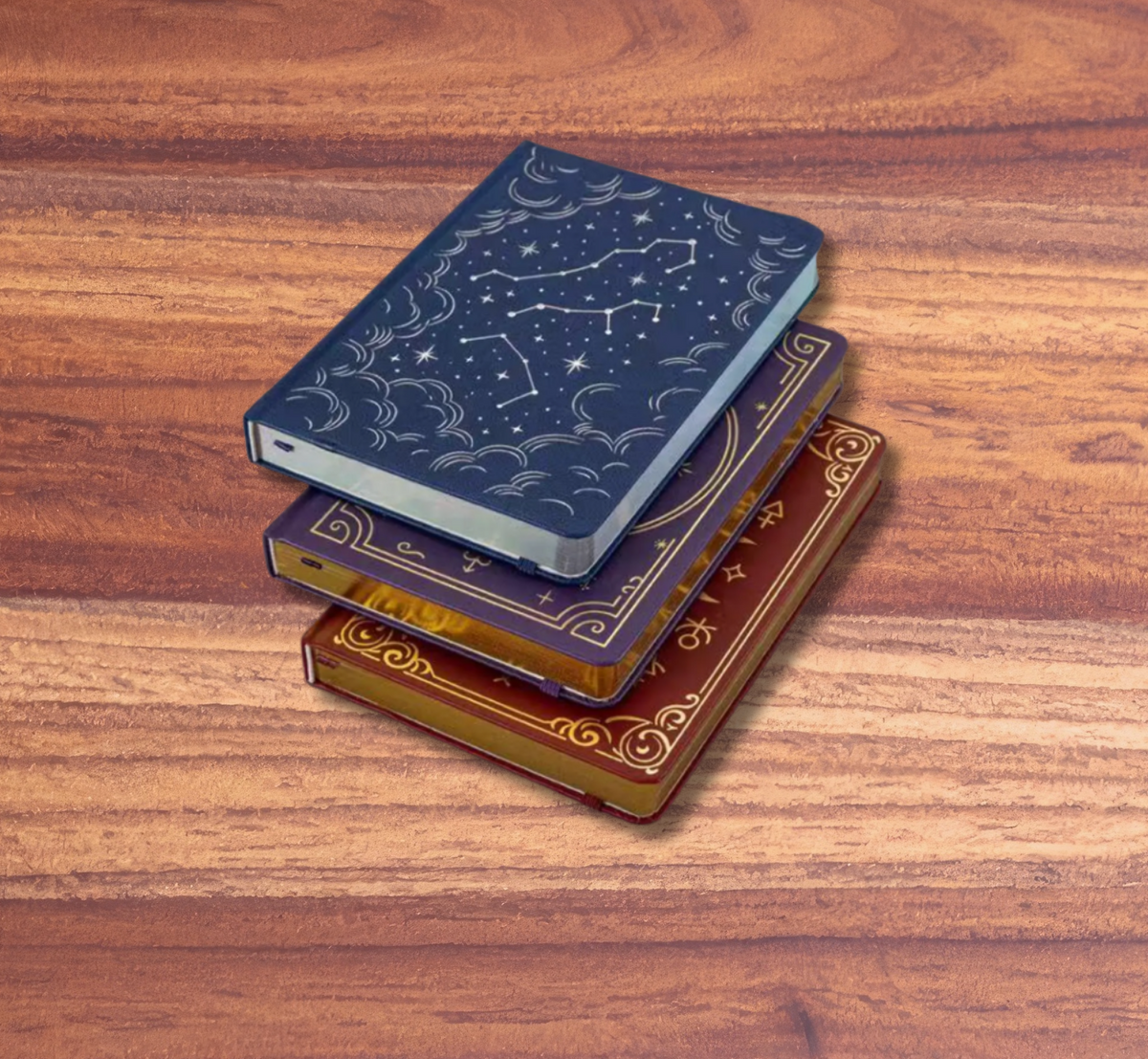 Three decorative hardcover notebooks stacked on a wooden tabletop, with a solid pink background. The top notebook is dark blue with an embossed starry constellation pattern, the middle is dark purple with gold borders, and the bottom is brown with gold ornate detailing.